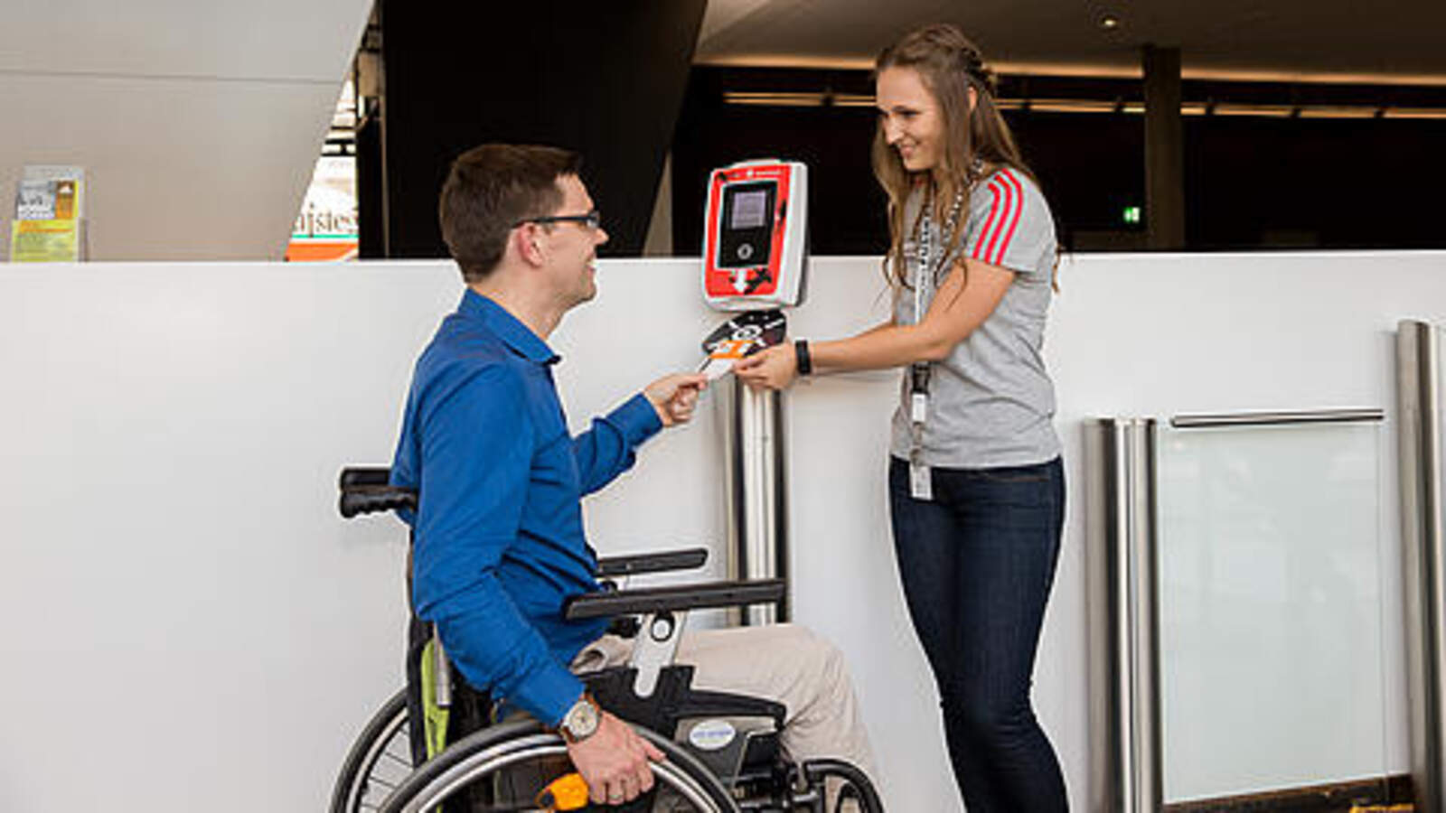 Ein Mann im Rollstuhl erhält Hilfe am Eingang: Die Mitarbeiterin zeigt ihm, wie er sein Ticket entwertet