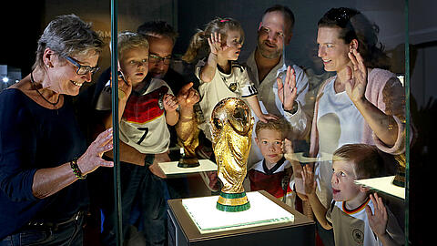 [Translate to en:] Eine große Familie steht um die Vitrine mit dem WM-Pokal herum