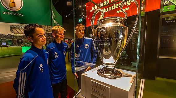 Jugendliche Fußballspieler in blauen Trainingsanzügen betrachten den Champions-League-Pokal im Deutschen Fußballmuseum in Dortmund.