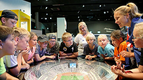 Kinder stehen im Kreis und lauschen gespannt den Worten einer Museumsführerin